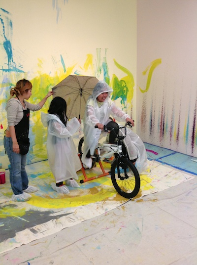 Students operate a stationary bicycle to create paint splatters on the wall.