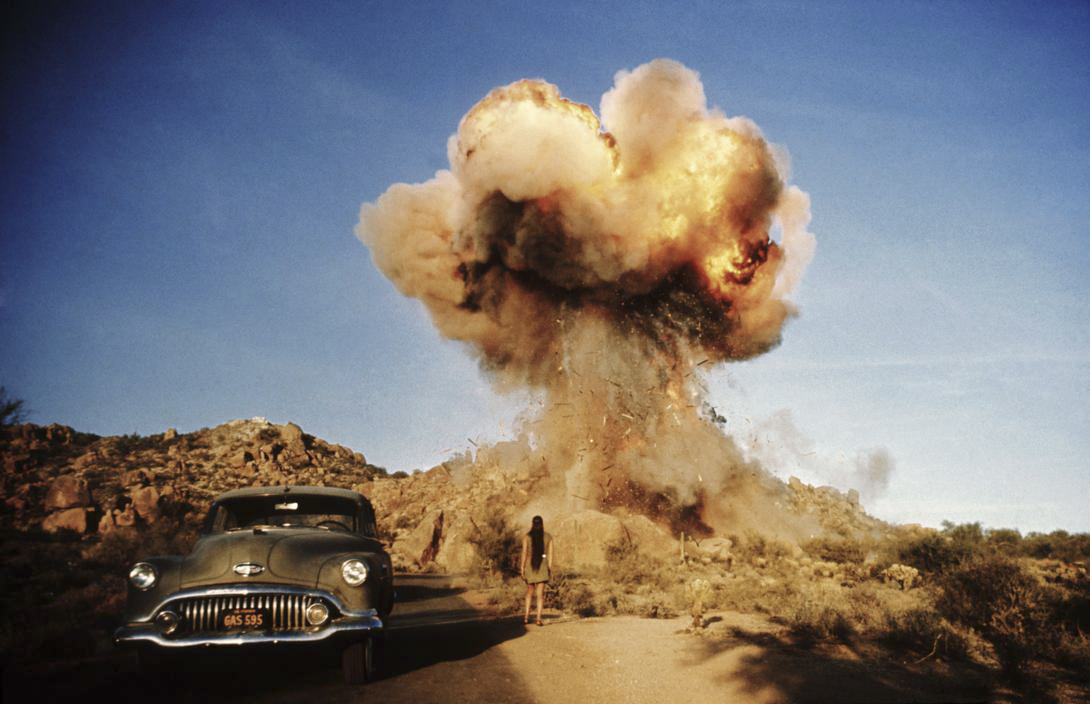 Woman standing on dirt road next to old car looking at an explosion