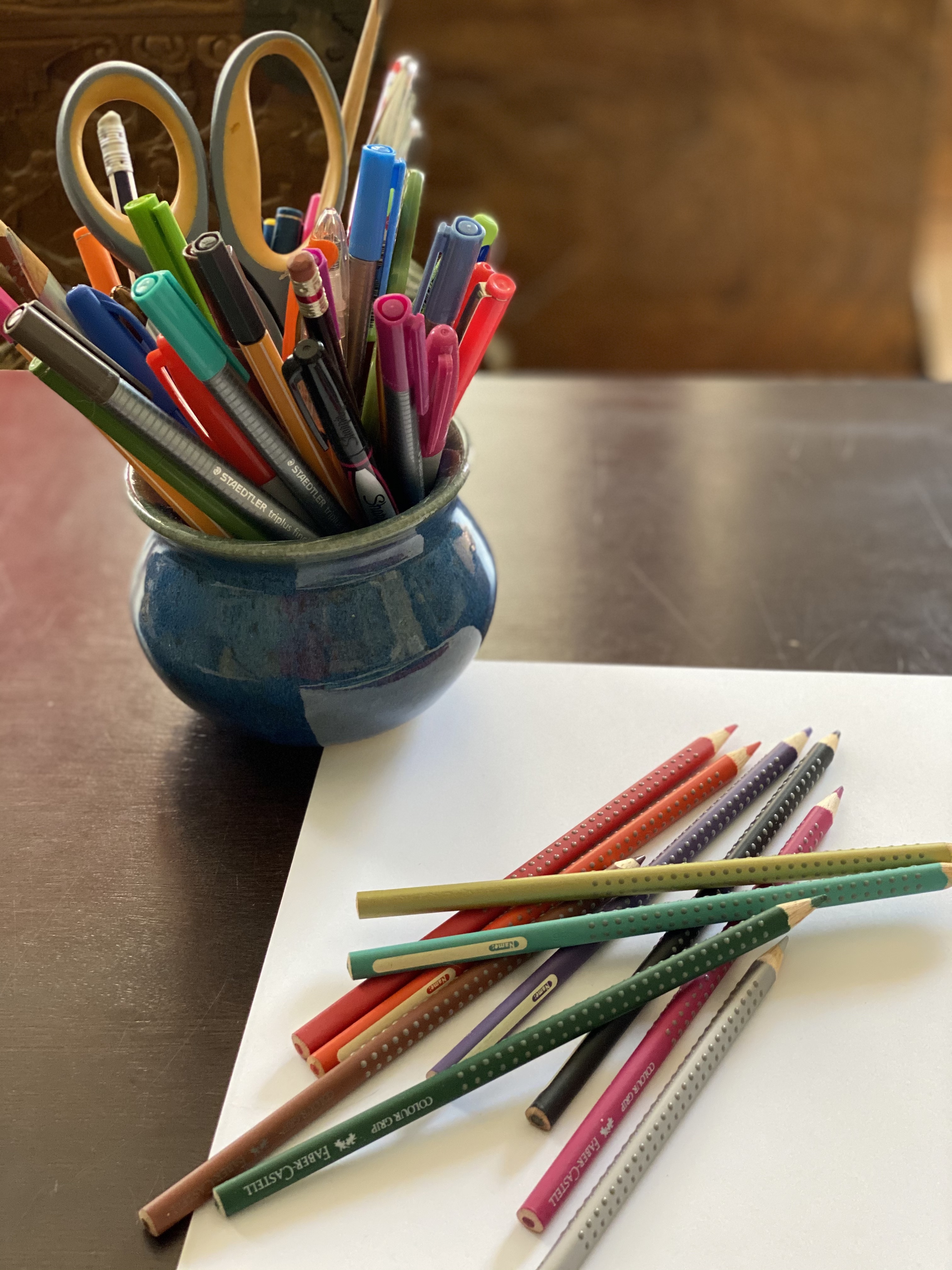 Ceramic pot holding pens and markers and scissors next to a sheet of paper and handful of colored pencils