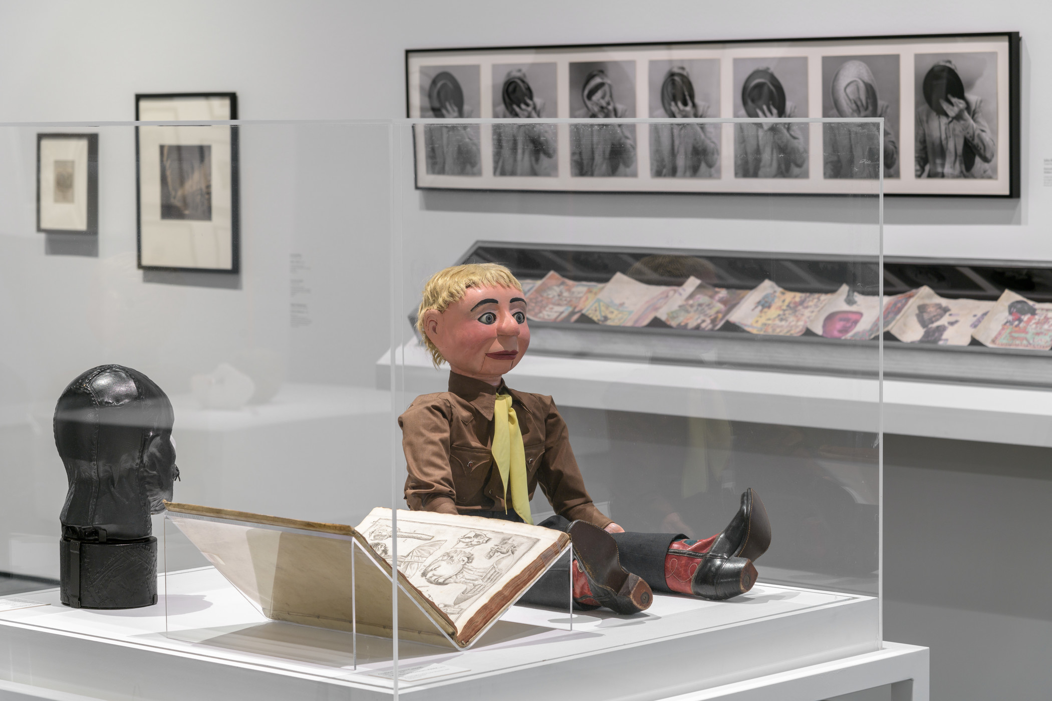 A ventriloquist dummy in a brown shirt, yellow tie, and red cowboy boots sits next to an open book and a black leather bust in a vitrine in a gallery, with framed photographs in the background 