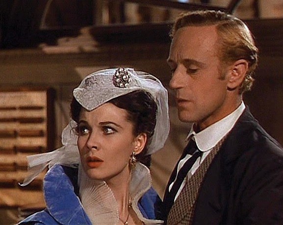 Still from the film Gone with the Wind with the actor wearing a white hat adorned with a sparkling white gem and white veil attached to the back, next to a man
