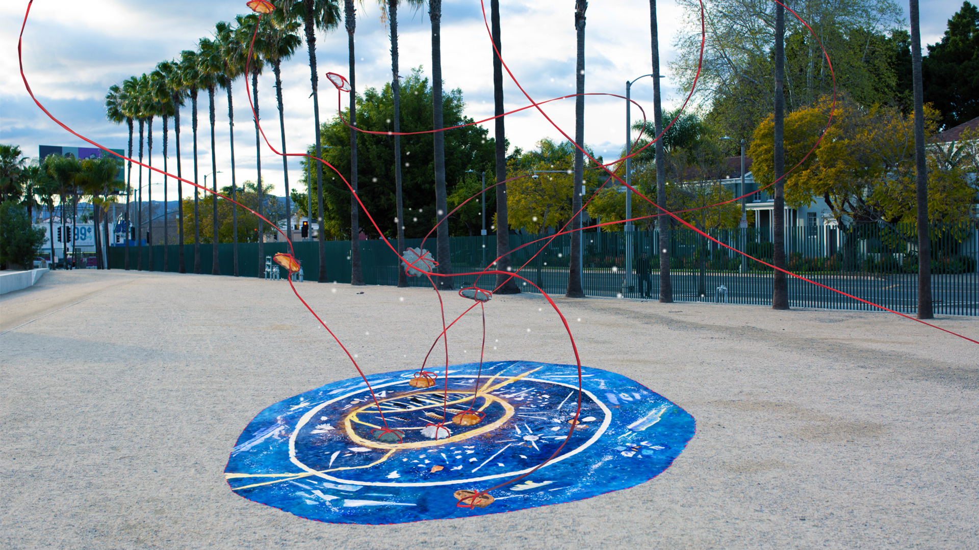 Red swirling lines reach into the sky from a round blue mat laid on decomposed granite with palm trees in the background
