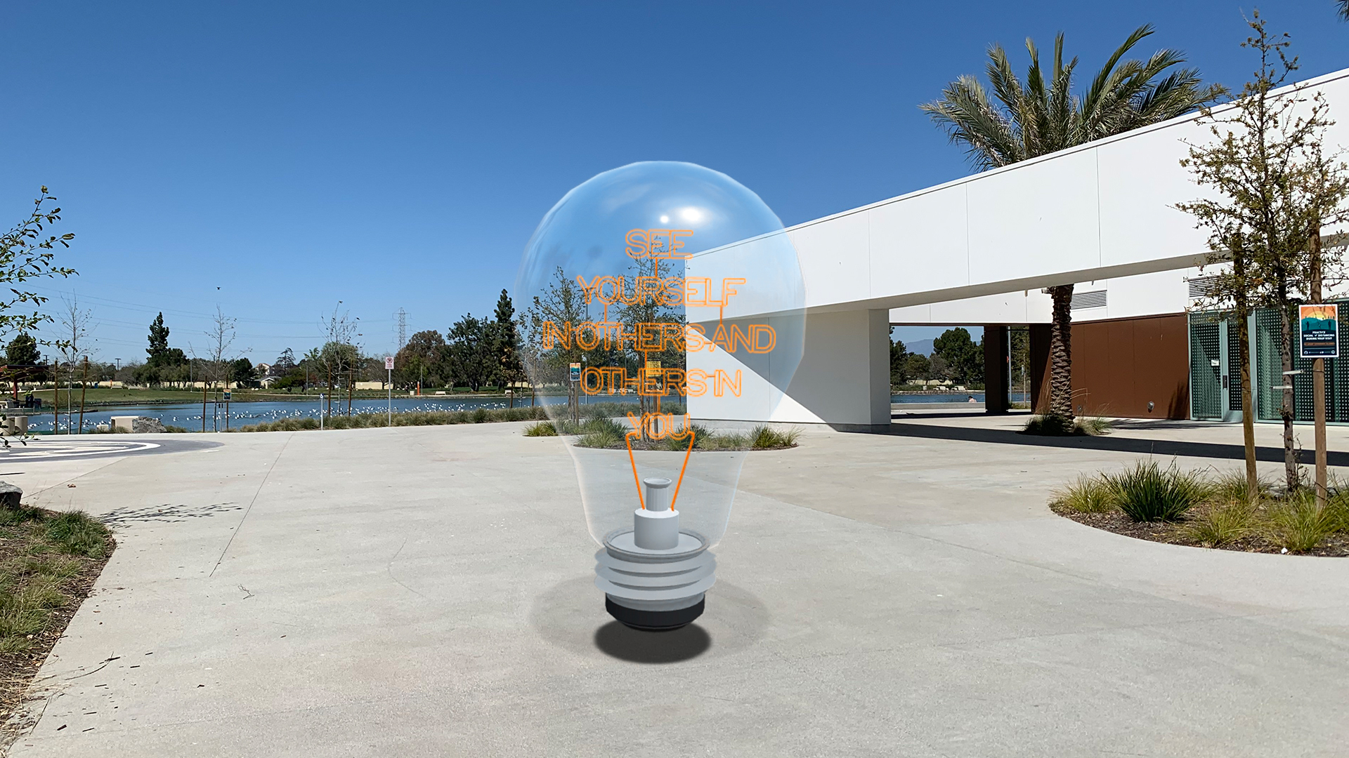 A giant lightbulb with writing on it floats on a concrete plaza with a building and a lake in the background