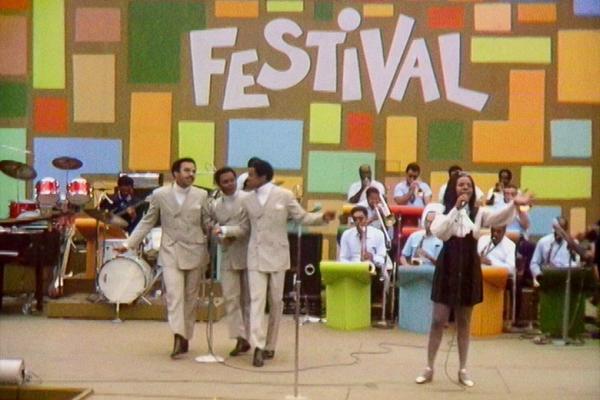 Film still of a stage with performers and the word "FESTIVAL" on a colorful backdrop