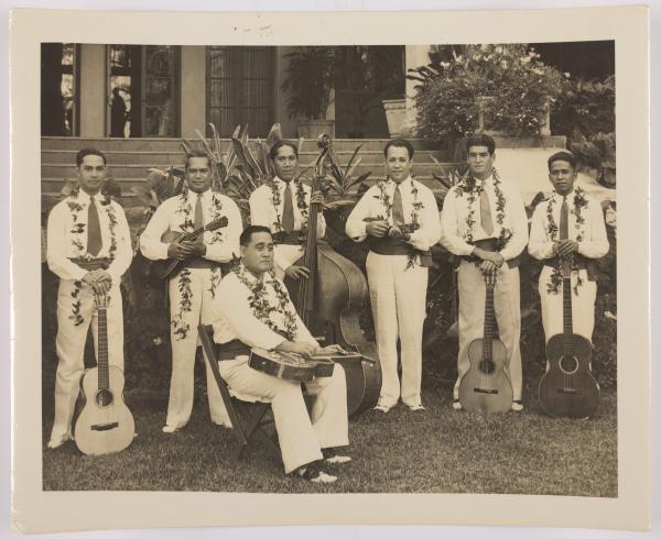 sepia toned image of a band standing outside a hotel—six men are standing, one is sitting in a chair in front of the group, all are wearing collared shirts, ties, dress pants