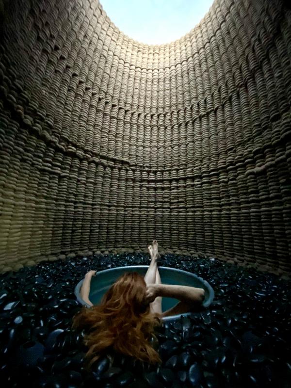 woman with red hair sitting in round tub that is submerged in black rocks inside a cylindrical-shaped structure, a circle of blue sky visible at top through opening in structure