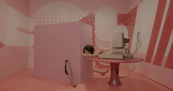 Woman's head and limbs emerge from a pink box to type on a computer in a surreal video still