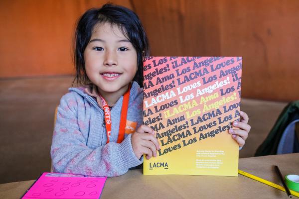 Photo of young girl holding a book that says "LACMA Loves Los Angeles!" on the cover
