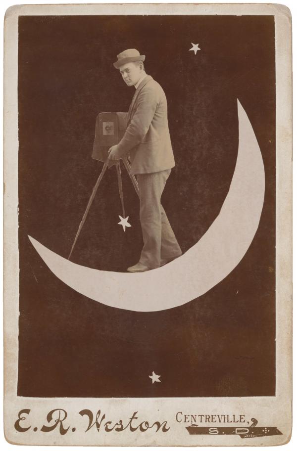 Black and white photo of a man on a cut-out moon with a camera and tripod