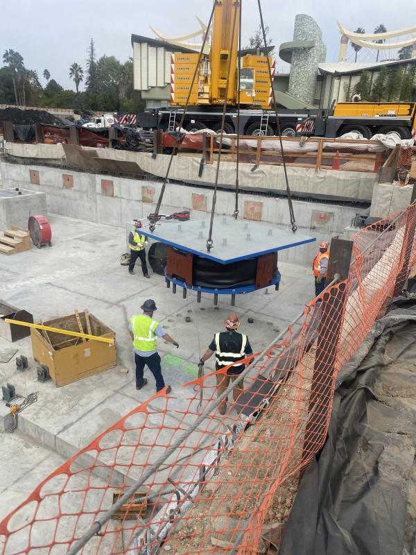 Four workers observing a large steel isolator being craned into construction site from overhead