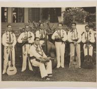 sepia toned image of a band standing outside a hotel—six men are standing, one is sitting in a chair in front of the group, all are wearing collared shirts, ties, dress pants
