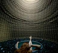 woman with red hair sitting in round tub that is submerged in black rocks inside a cylindrical-shaped structure, a circle of blue sky visible at top through opening in structure