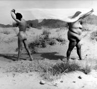 black and white image of two nude women standing apart, facing away from each other in the desert, a sheer piece of fabric stretched out between them, held over their heads