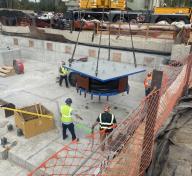 Four workers observing a large steel isolator being craned into construction site from overhead