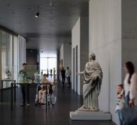 Visitors in light and art-filled gallery with floor-to-ceiling windows on the left side of image
