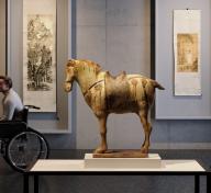 A rendering of a gallery with an ancient horse sculpture in the center and two ink painting scrolls hanging on the wall in the background. On the left is a man in a wheelchair and on the right a figure walking into the gallery.